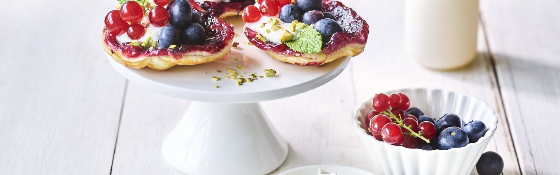 Upside down taartje met aalbessen en blauwe bessen Op een witte taartschaal liggen 3 kleine taartjes met zomerfruit. Hiernaast ligt nog wat opgeklopte room, nog een schaaltje met extra rood fruit en een vaasje met roze bloemetjes.
