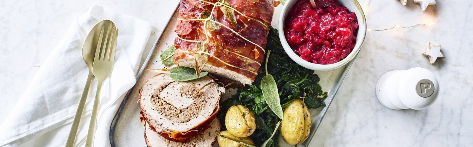 Varkensrollade met gehakt, veenbessencompote en krieltjes uit de oven Een mooi aangesneden varkensrollade ligt op een rechthoekig dienblad. De aardappeltjes werden net niet doormidden gesneden alvorens ze de oven in gingen met een kruidentakje. De veenbessencompote staat langs de rollade die zorgt voor een intense kleur in deze feestmaaltijd. Goud bestek ligt langs het gerecht.