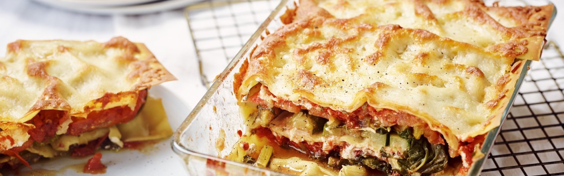 Vegetarische lasagne met snijbiet, courgette en ricotta De lasagne staat op tafel in een glazen doorzichtige ovenschotel. Hij is aangesneden zodat je mooi de laagjes ziet van pasta, groenten en saus. De portie die is uitgeschept ligt op een bordje er naast.