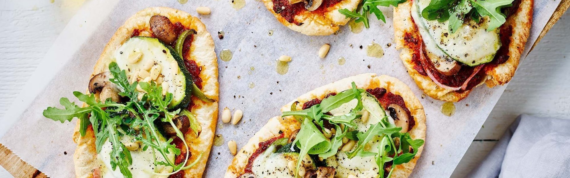 Vier kleine pizza's liggen op een houten plank, klaar om aan te vallen. De pizzabodems zijn pitabroodjes: snel, simpel en lekker. De pizza's zijn belegd met tomatensaus, geroosterde groenten en kaas. De rucola werd achteraf toegevoegd aan de pizza. 