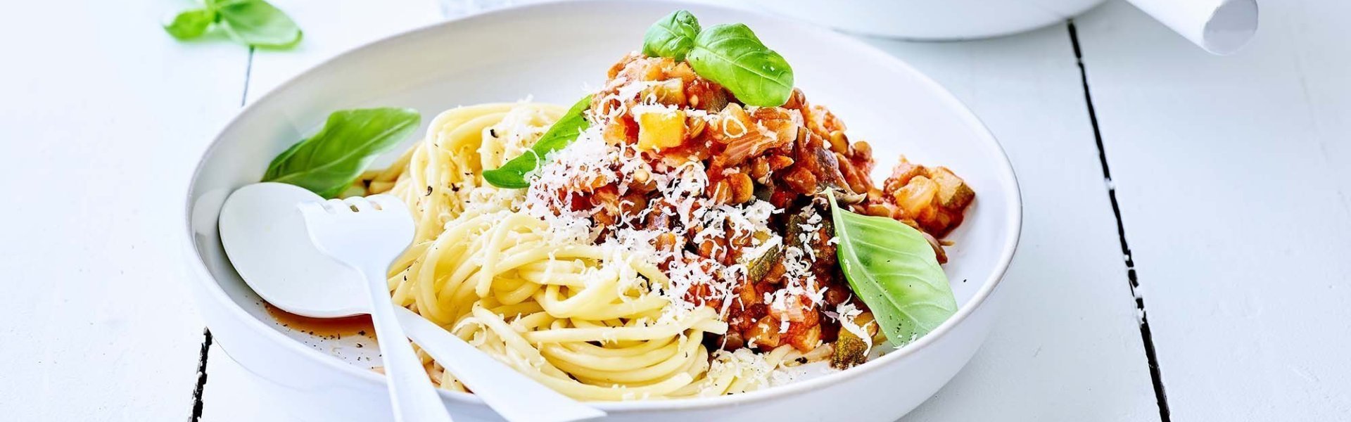 Spaghetti geserveerd in wit bord met portie saus half over de pasta. Versgemalen kaas over de saus met enkele verse basilicumblaadjes. 