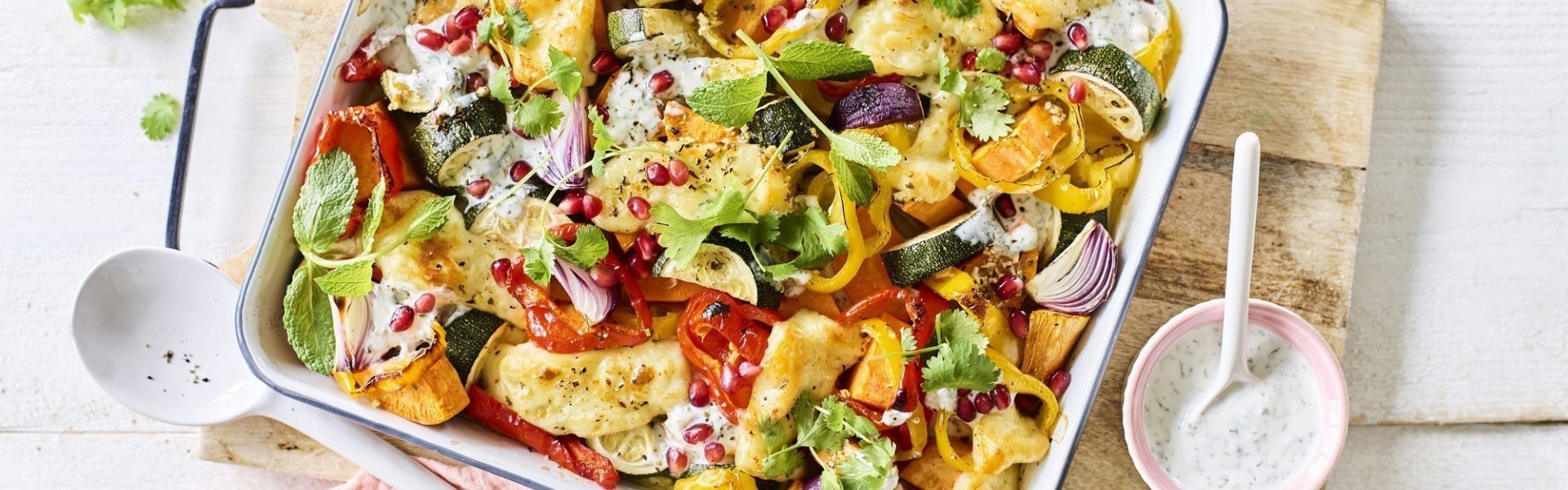 Vegetarische traybake met aardappel, rode ui, paprika en Berloumi Op een witte tafel staat een houten snijplank met hierop een vierkante ovenschotel. Hij is gevuld met zoete aardappel, courgette, paprika en rode ui. Er is ook Berloumi gebruikt in dit gerechtje.