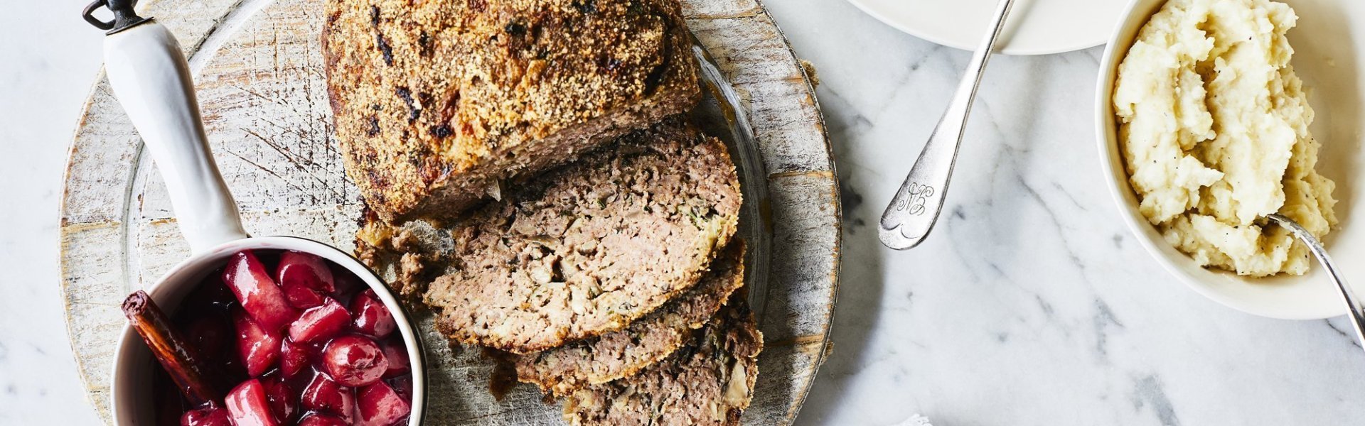 Vleesbrood Een vleesbroodje ligt aangesneden op een snijplank met een potje heerlijke warme krieken naast. Er staat een bordje langs die gevuld is met deze maaltijd.