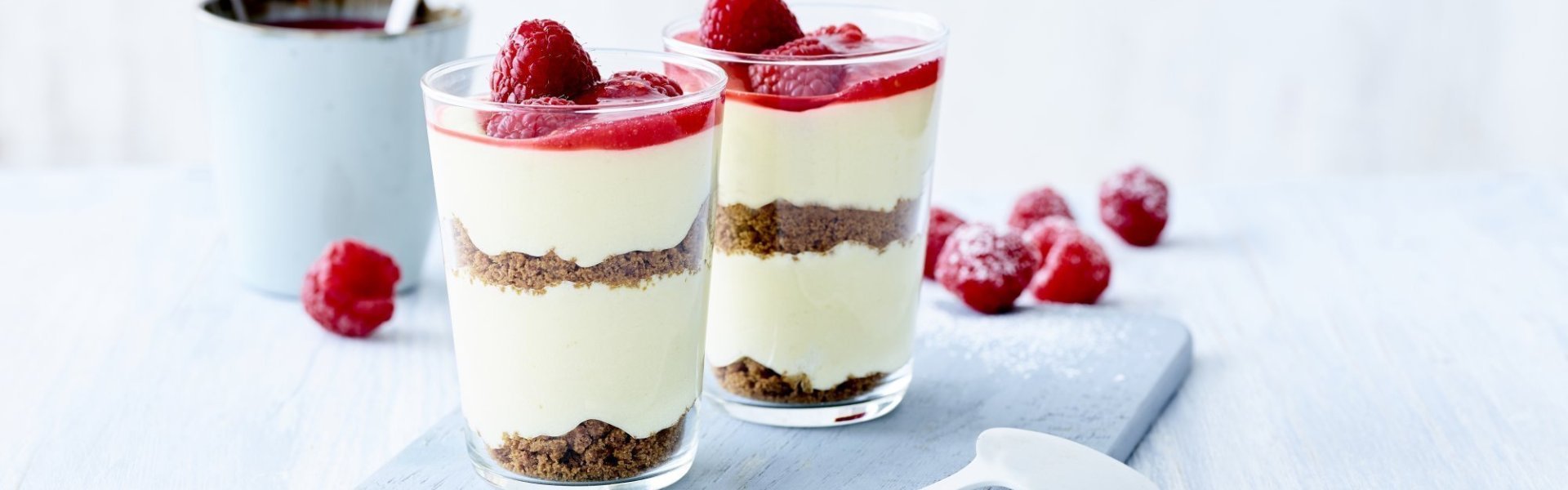 Witte chocolademousse met laagjes speculaas en bovenop afgewerkt met een coulis van frambozen. Ze zijn geserveerd in hoge glazen, waardoor je instant een feestlook krijgt. Tas koffie staat links naast de chocolademousse en enkele frambozen liggen op de tafel.