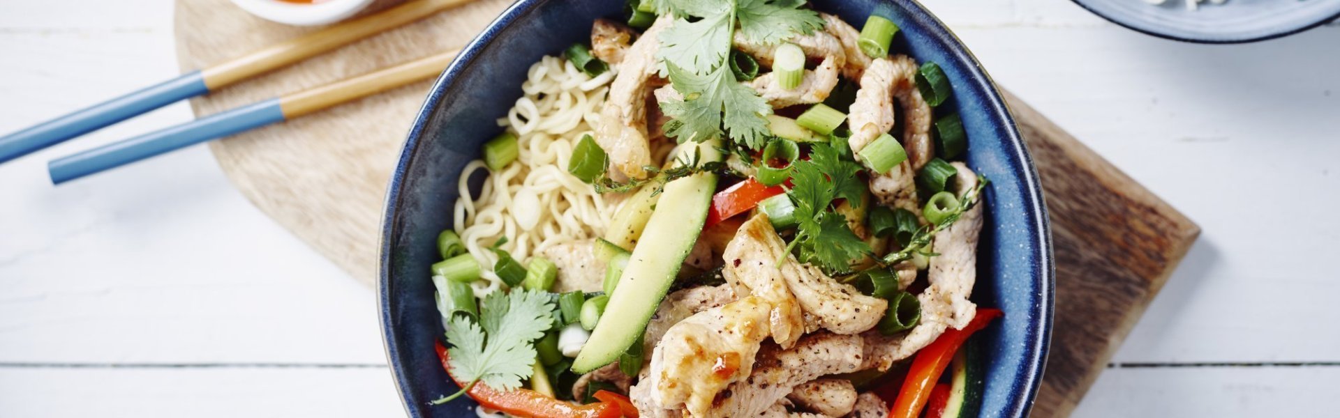 Donker blauwl bord gevuld met een wok van varkensreepjes, oesterzwammen, courgette en paprika. Bij dit bordje liggen eetstokjes, een potje loempiasaus en verse noodles.