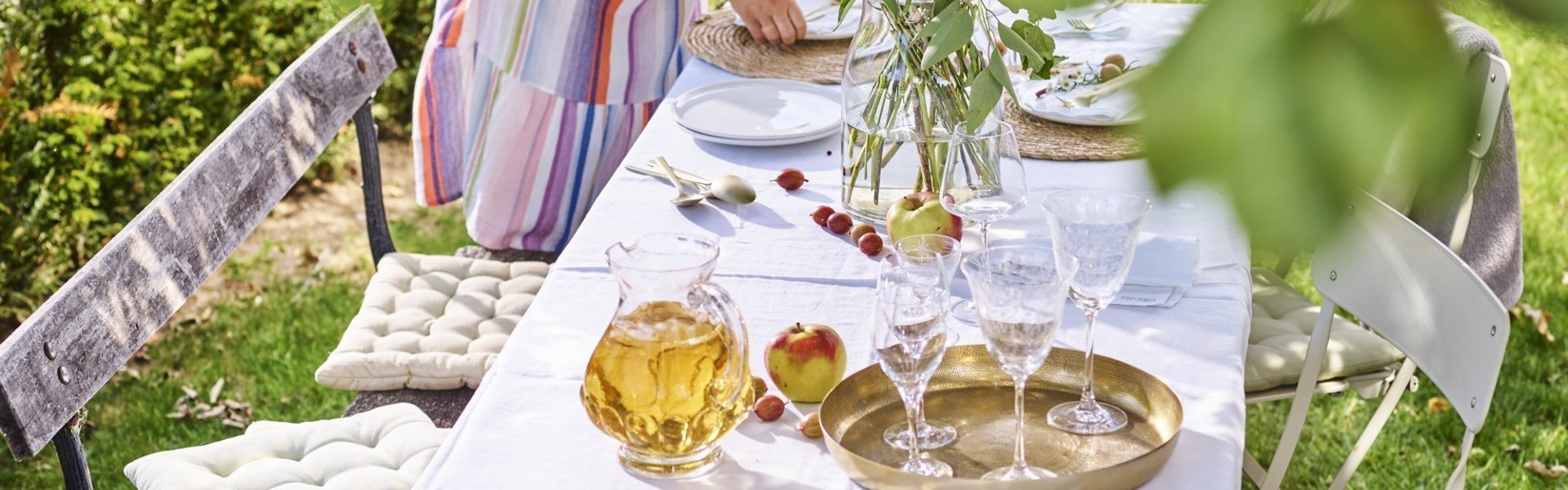 Dekken van zomertafel Vrouw met gestreept zomerjurk is de tafel aan klaarzetten voor enkele gasten. Er staat al een kan met vers appelsap op tafel en enkele glazen. De vrouw is de borden aan het plaatsen op de tafel met wit linnen tafelkleed. De tafel staat onder een perenboom in de tuin.