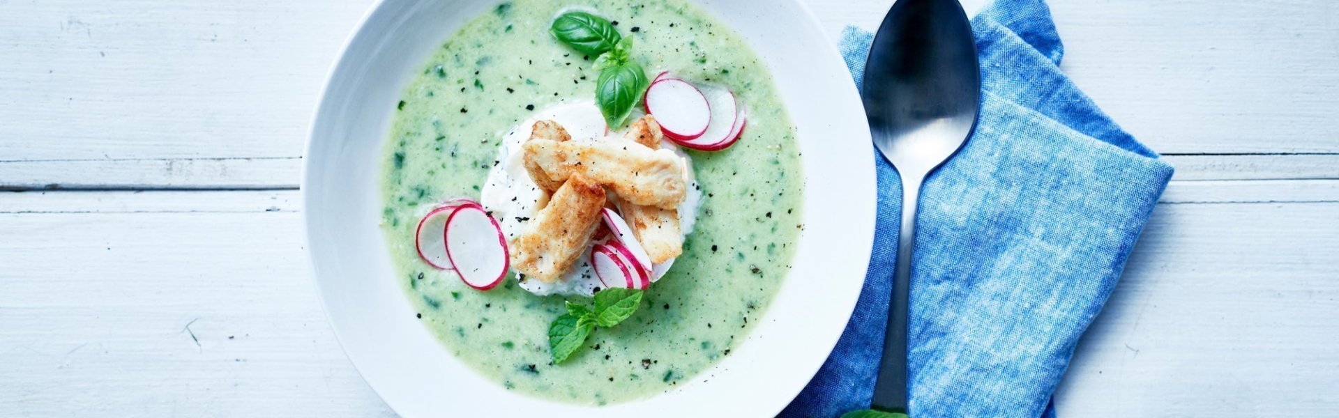 gazpacho met krokante tongscharreepjes Een gazpacho op basis van komkommer, avocado en verse kruiden met een slaatje van appel en selder in het midden van het bord. Hierop liggen de tonscharreepjes met schijfjes radijs en verse kruiden. Er ligt een zilveren lepel naast met een linnen blauwe servette.