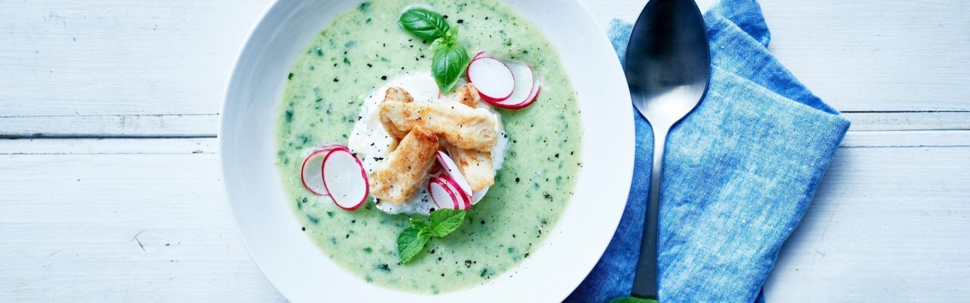 Een gazpacho op basis van komkommer, avocado en verse kruiden met een slaatje van appel en selder in het midden van het bord. Hierop liggen de tonscharreepjes met schijfjes radijs en verse kruiden. Er ligt een zilveren lepel naast met een linnen blauwe servette.