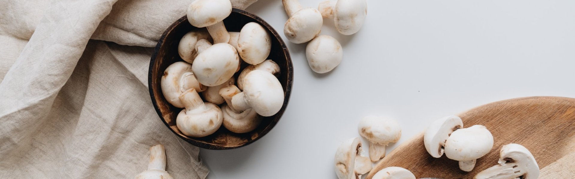 Parijse champignons op wit aanrecht. Enkele champignons zijn versneden in schijfjes op houten snijpland. Er licht een mes en linnen handdoek naast.