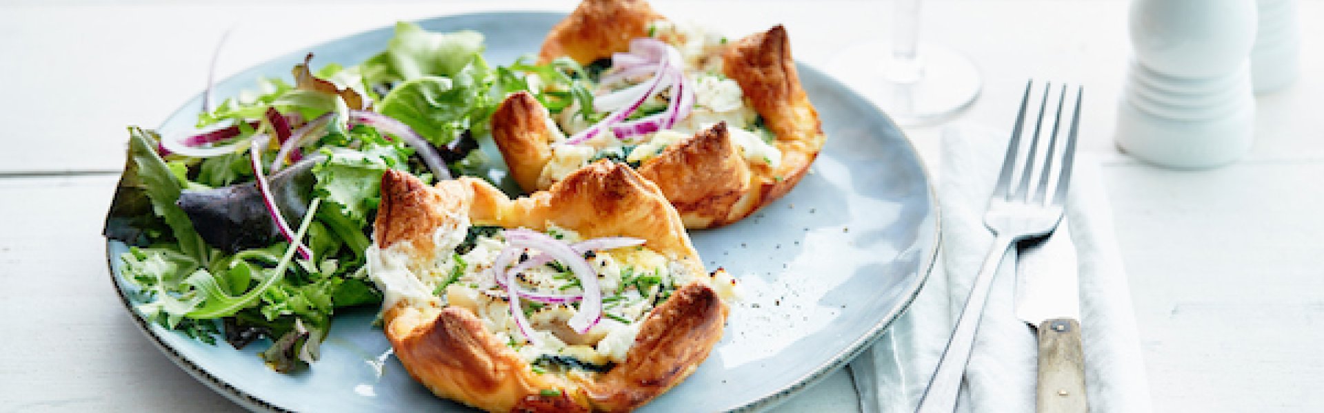 Bladerdeegtaartjes met vis van bij ons. Een quiche van steenbolk met spinazie en geitenkaas. Alles gepresenteerd op een blauwgrijs bordje met een gemengde groene salade en verse rode uiringen. Glaasje witte wijn staat rechtsbovenaan en een witte zoutmolen.