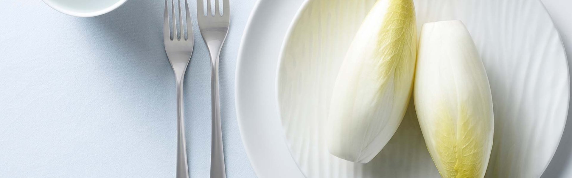 2 stronken witloof liggen op een bord. Vork en glas staan naast het bord, op tafel ligt ook een wit tafelkleed.